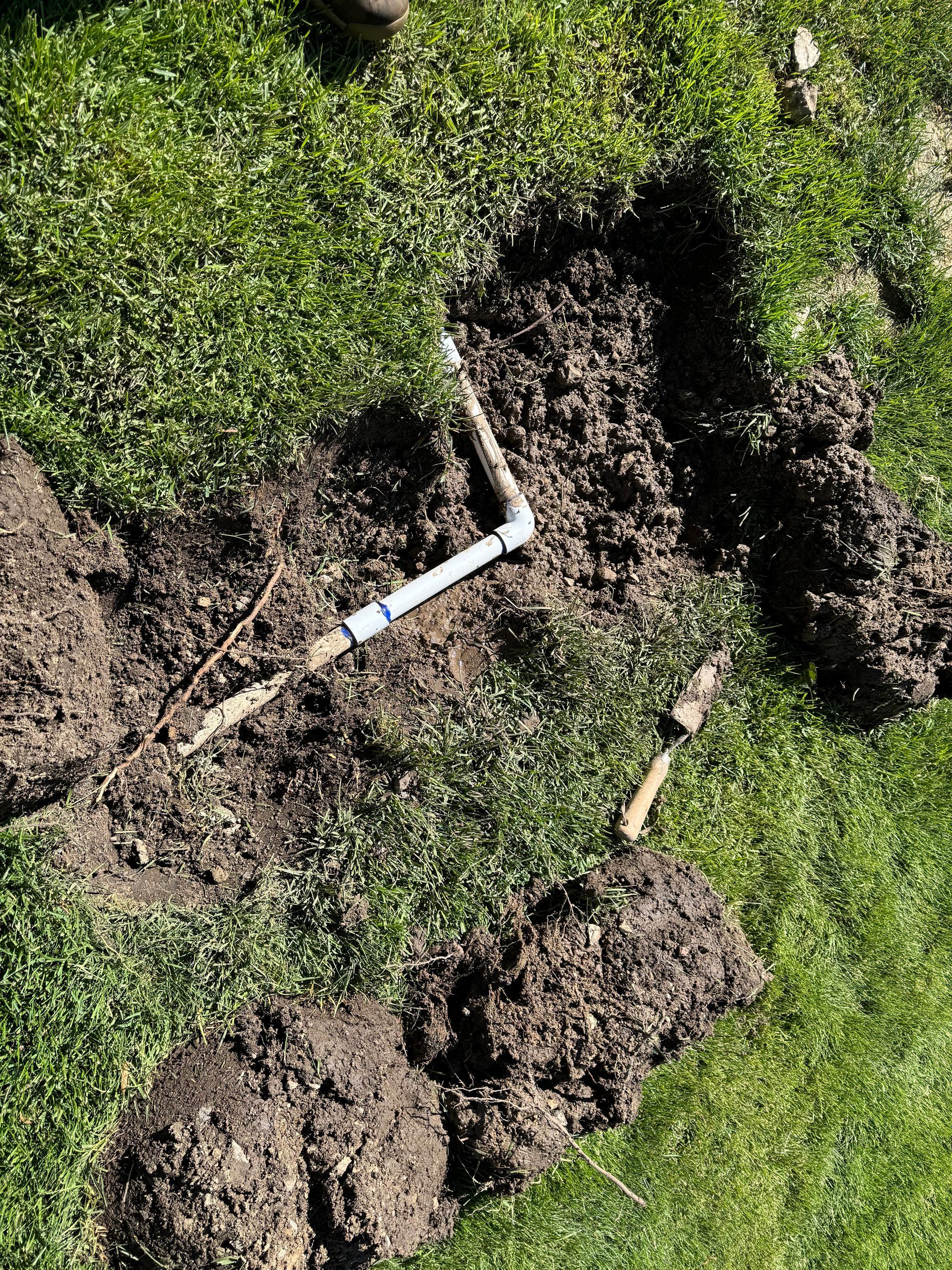 An exposed L-shaped PVC pipe connection in a hole dug in a grassy lawn.