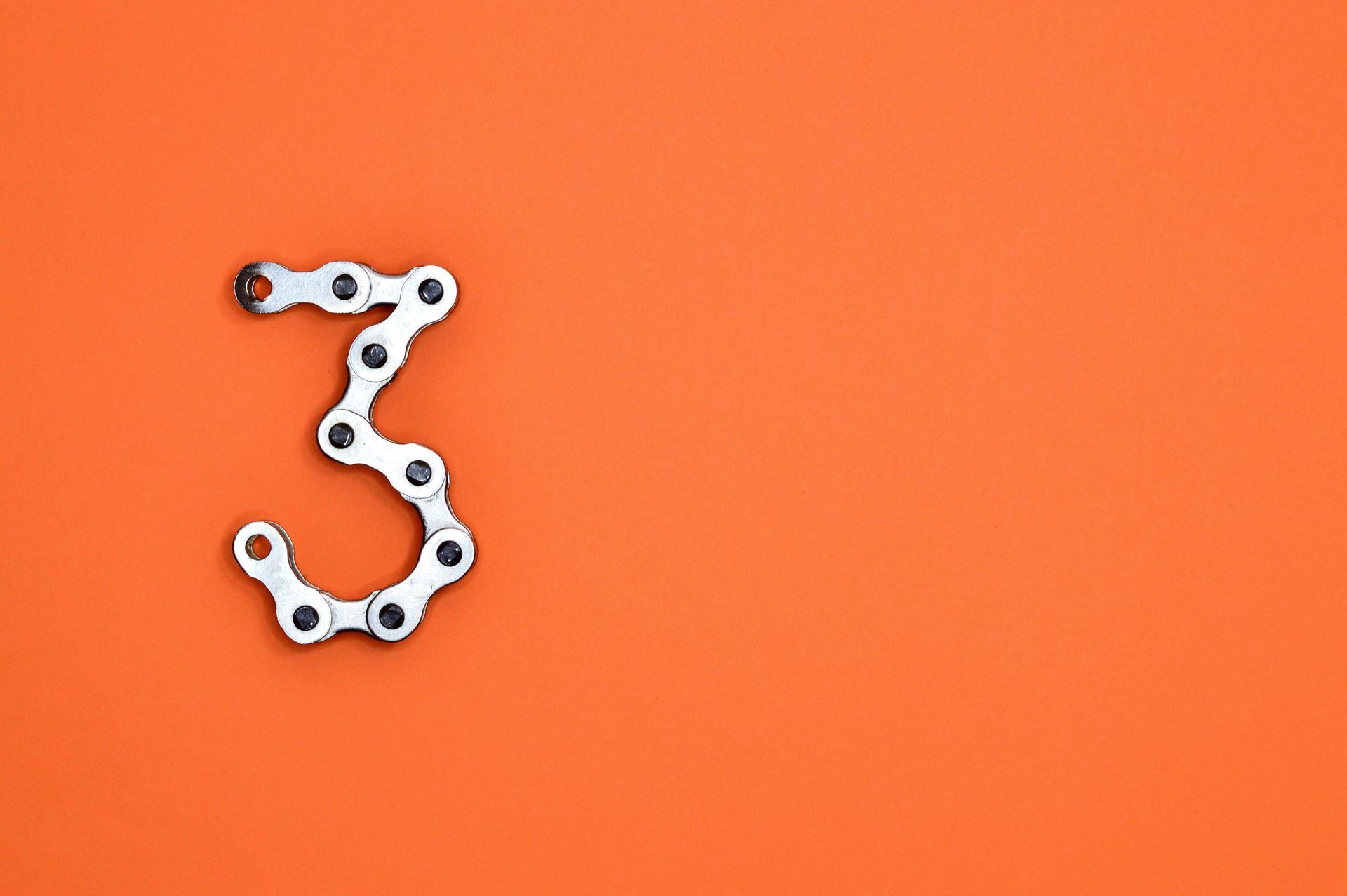 The number three formed from a silver metal bicycle chain on a solid orange background.