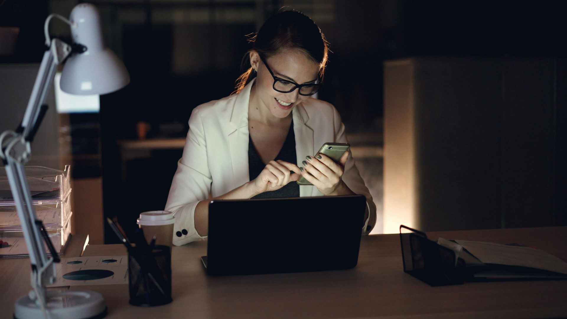 A person sits at a dimly lit desk working on a smartphone while a laptop is open in front of them.