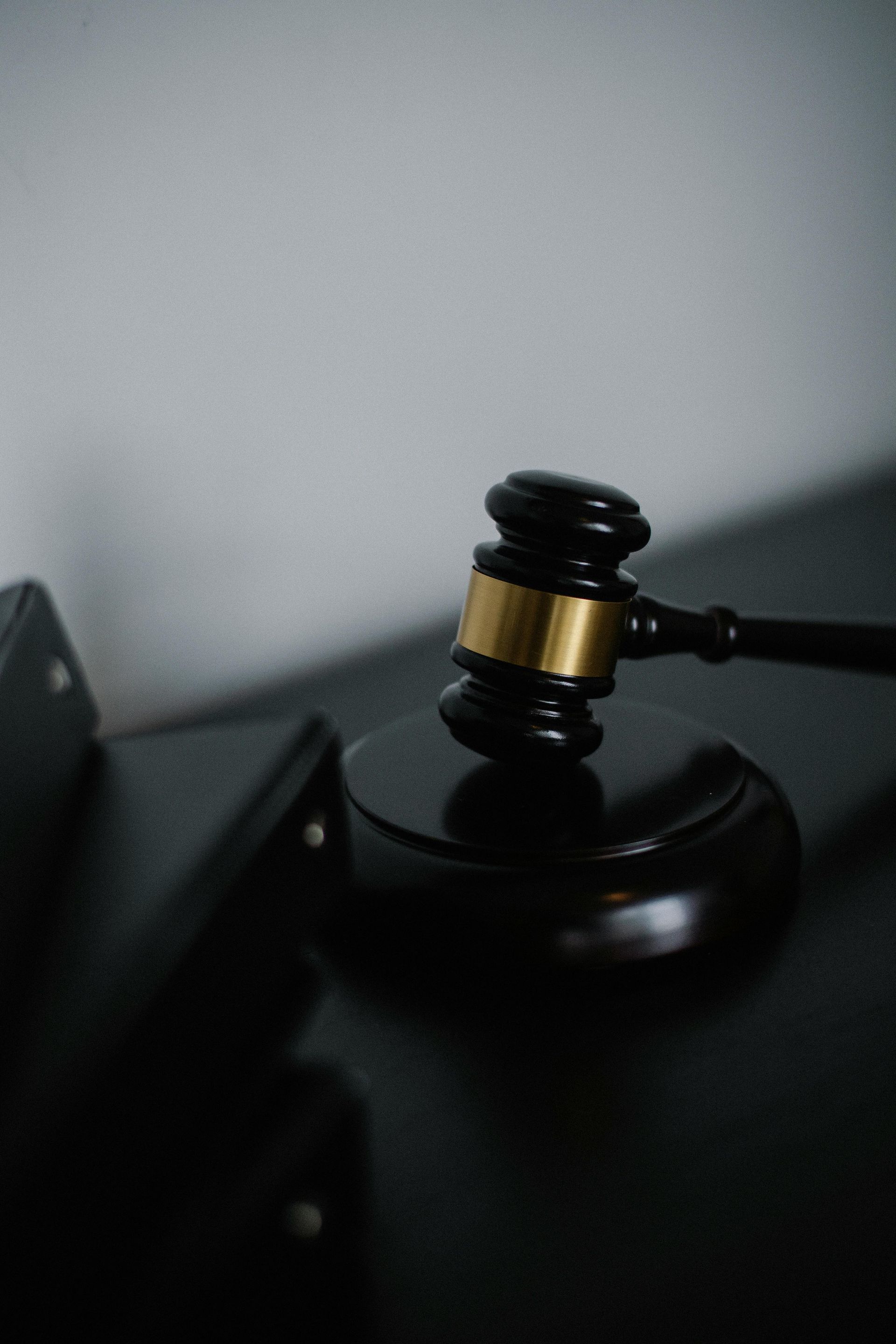 A black wooden judge's gavel with a gold band resting on a matching round base against a neutral gray background.