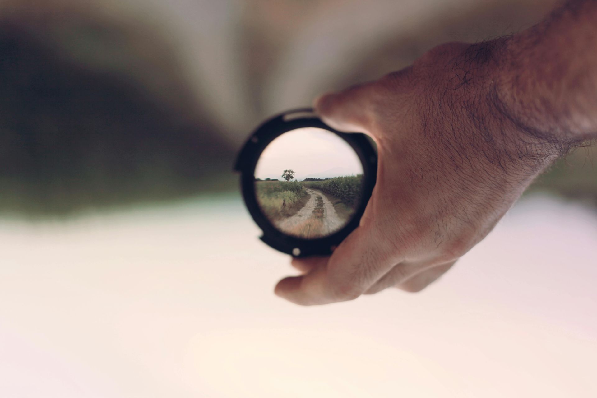 A hand holds a circular lens, focusing on a dirt path stretching through a field toward a distant tree.