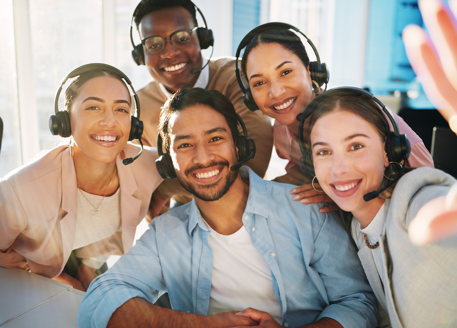 Five diverse coworkers wearing headsets smile while taking a group selfie in an office.