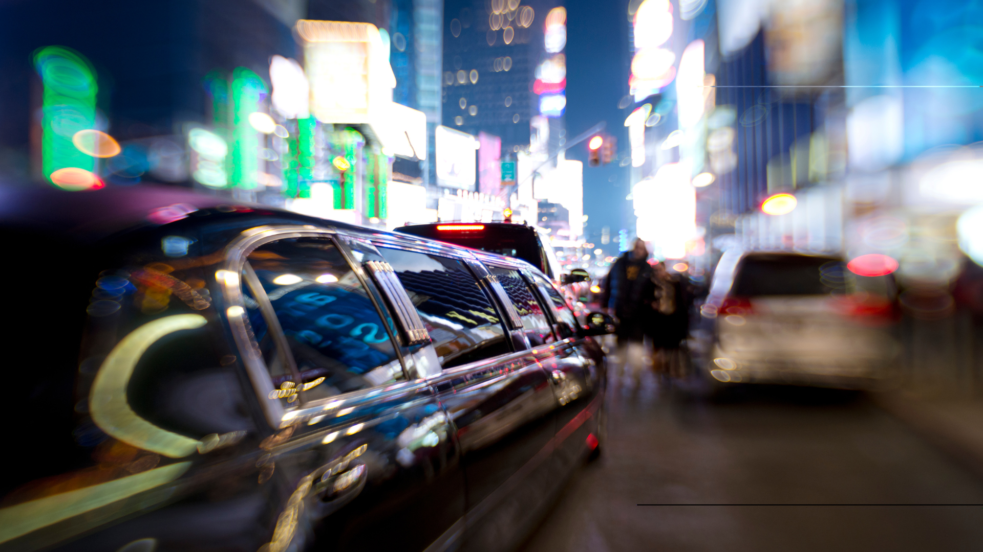A black limousine drives through a bright, blurry Times Square at night.