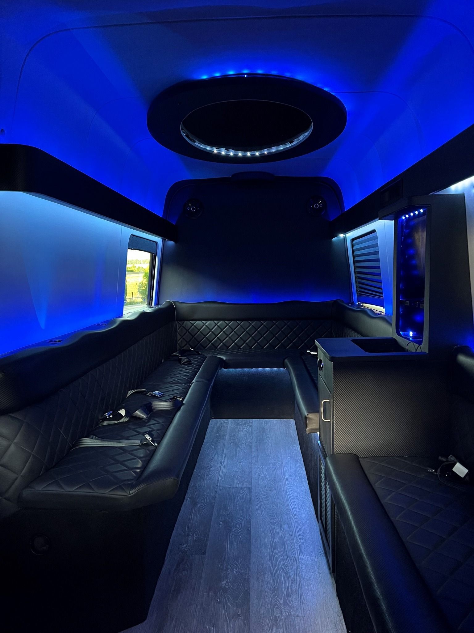 Interior of a limousine with blue lights. Black seating with diamond stitching, dark ceiling, and a central light fixture.