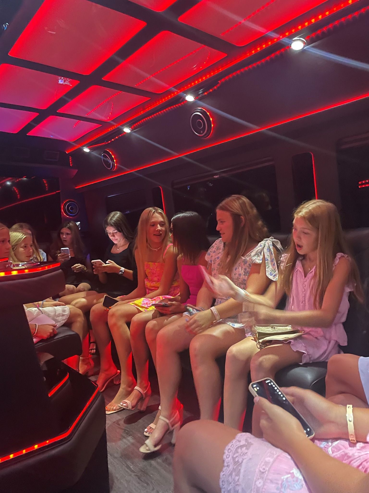 Inside a red-lit party bus, girls in dresses are seated, using phones.