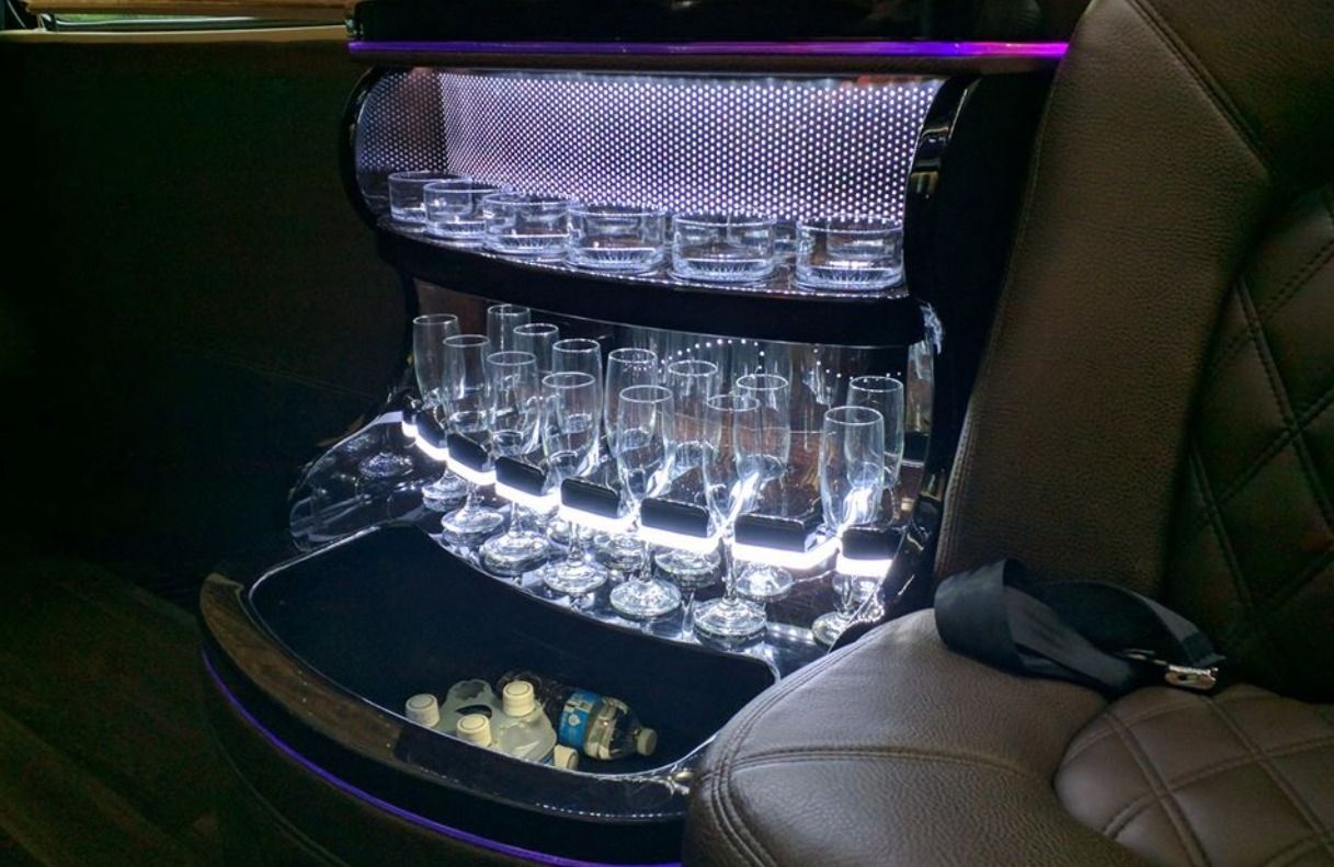 Interior of a limousine. Illuminated bar with glasses and ice bucket, dark upholstery on the right.