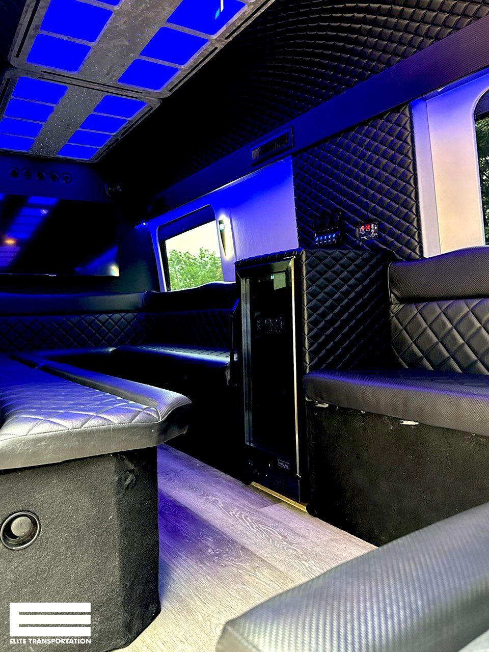 Interior of a limousine. Black quilted seats and walls, blue lighting. A refrigerator and window are visible.