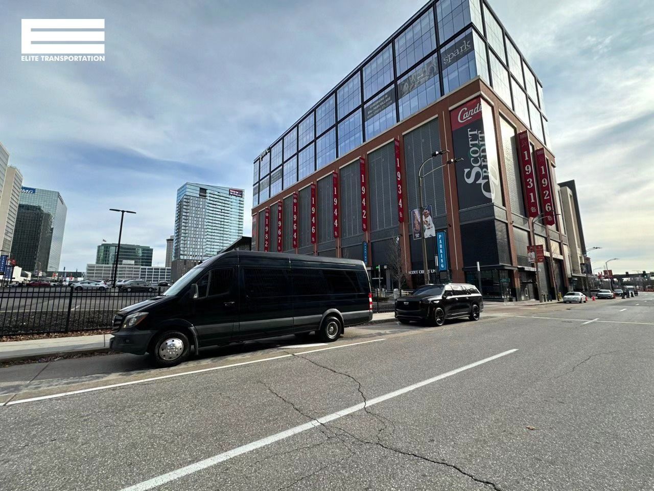 Black van and SUV parked on a city street, large building in the background.