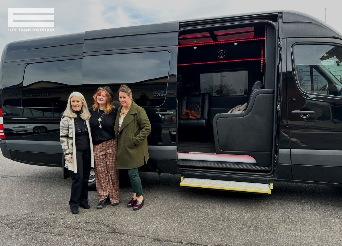 Three women pose next to a black van with an open door, revealing interior seating.