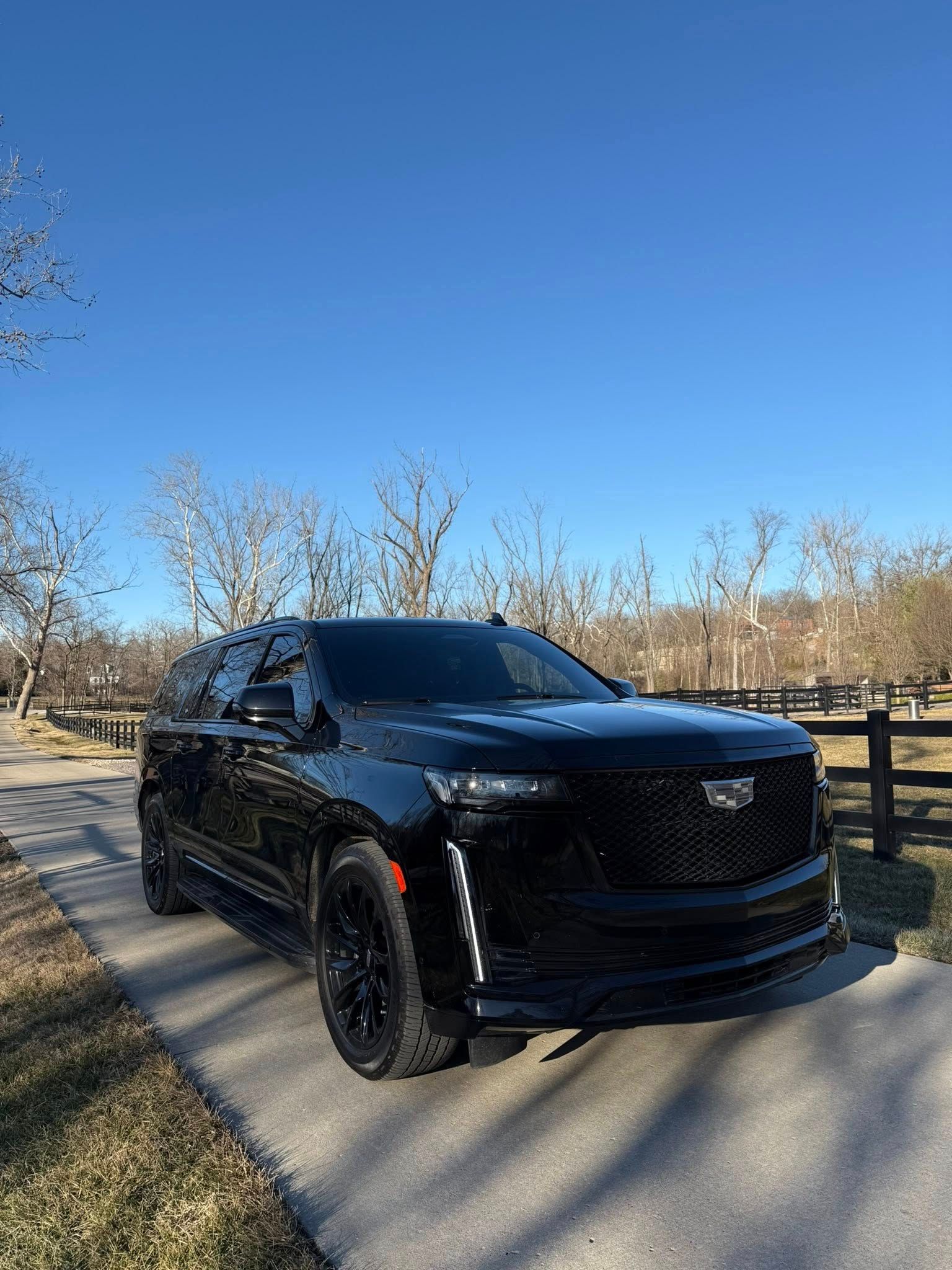 Black Cadillac Escalade parked on a paved pathway with a fence and bare trees in the background under a blue sky.