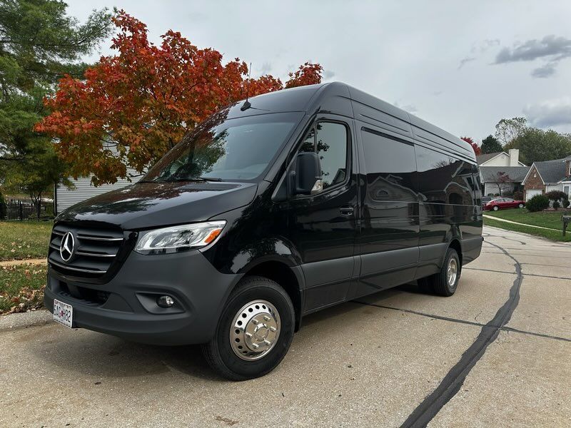 Black Mercedes Sprinter van parked on a suburban street beside a tree with autumn leaves