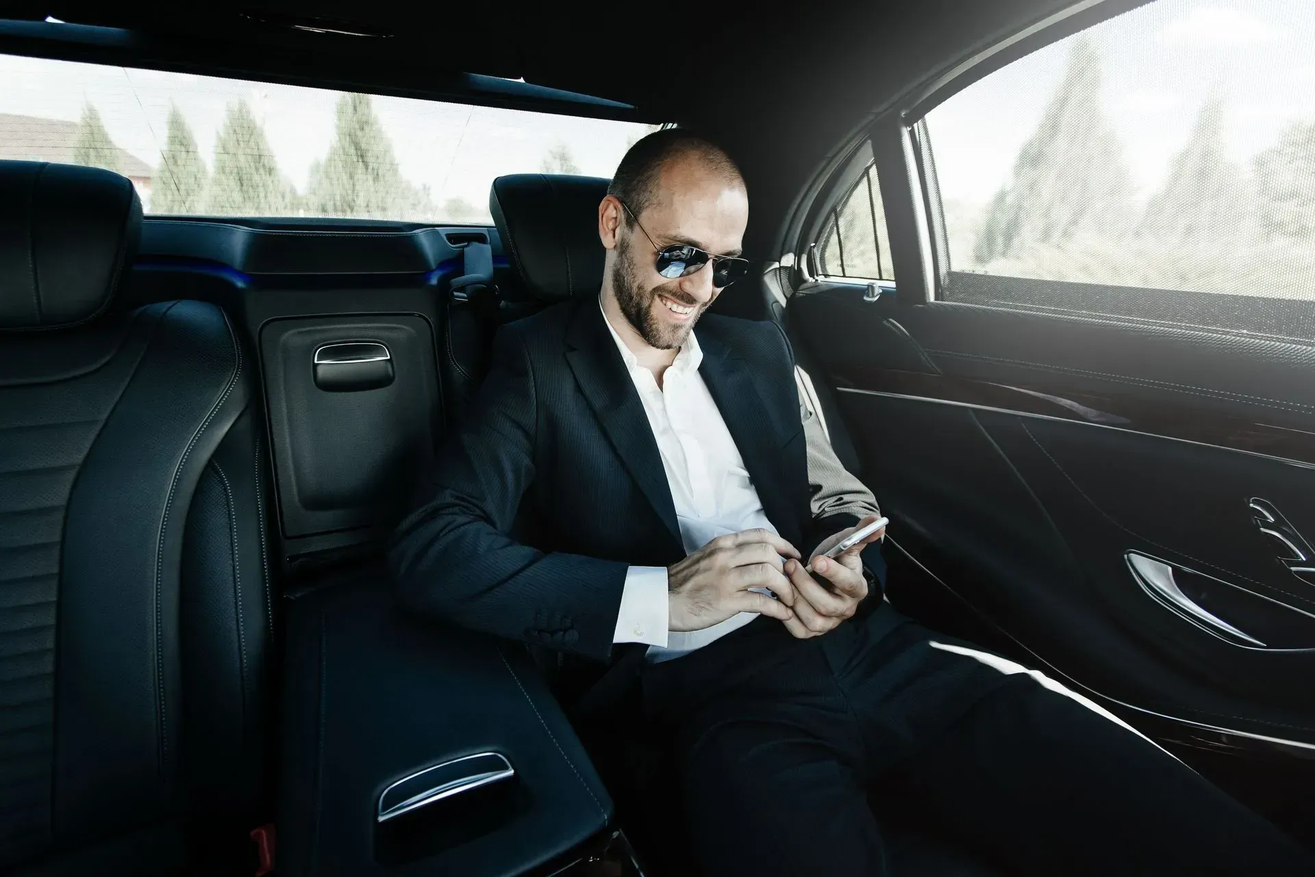 Man in suit and sunglasses smiles while looking at phone in the backseat of a car.