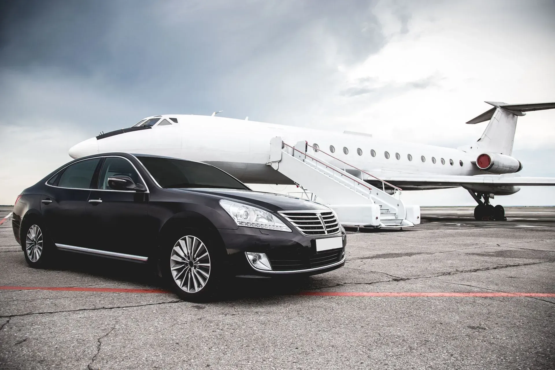 Dark sedan parked on tarmac, next to a white private jet with extended stairs; overcast sky.