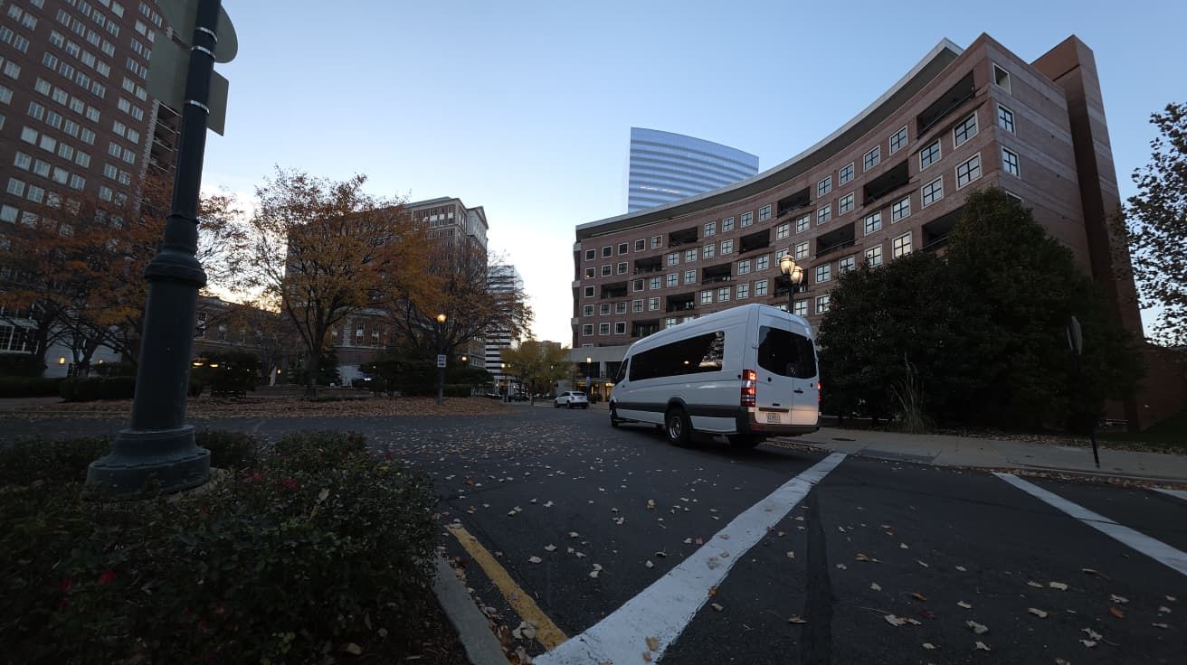 White van turning on a street with buildings and trees under a dusky sky.