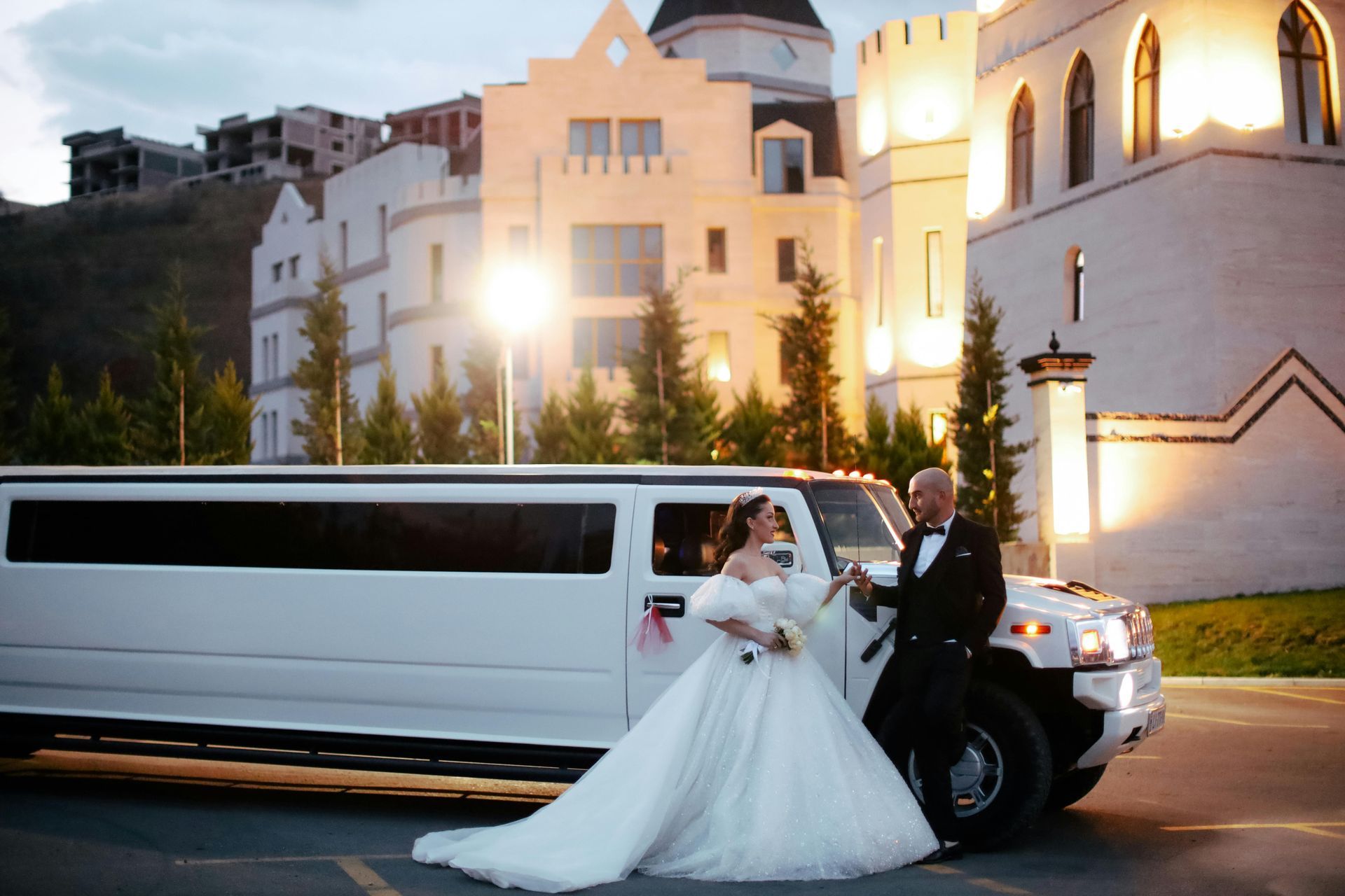 Bride and groom with a white limousine outside a castle-like building; the couple is toasting with glasses.