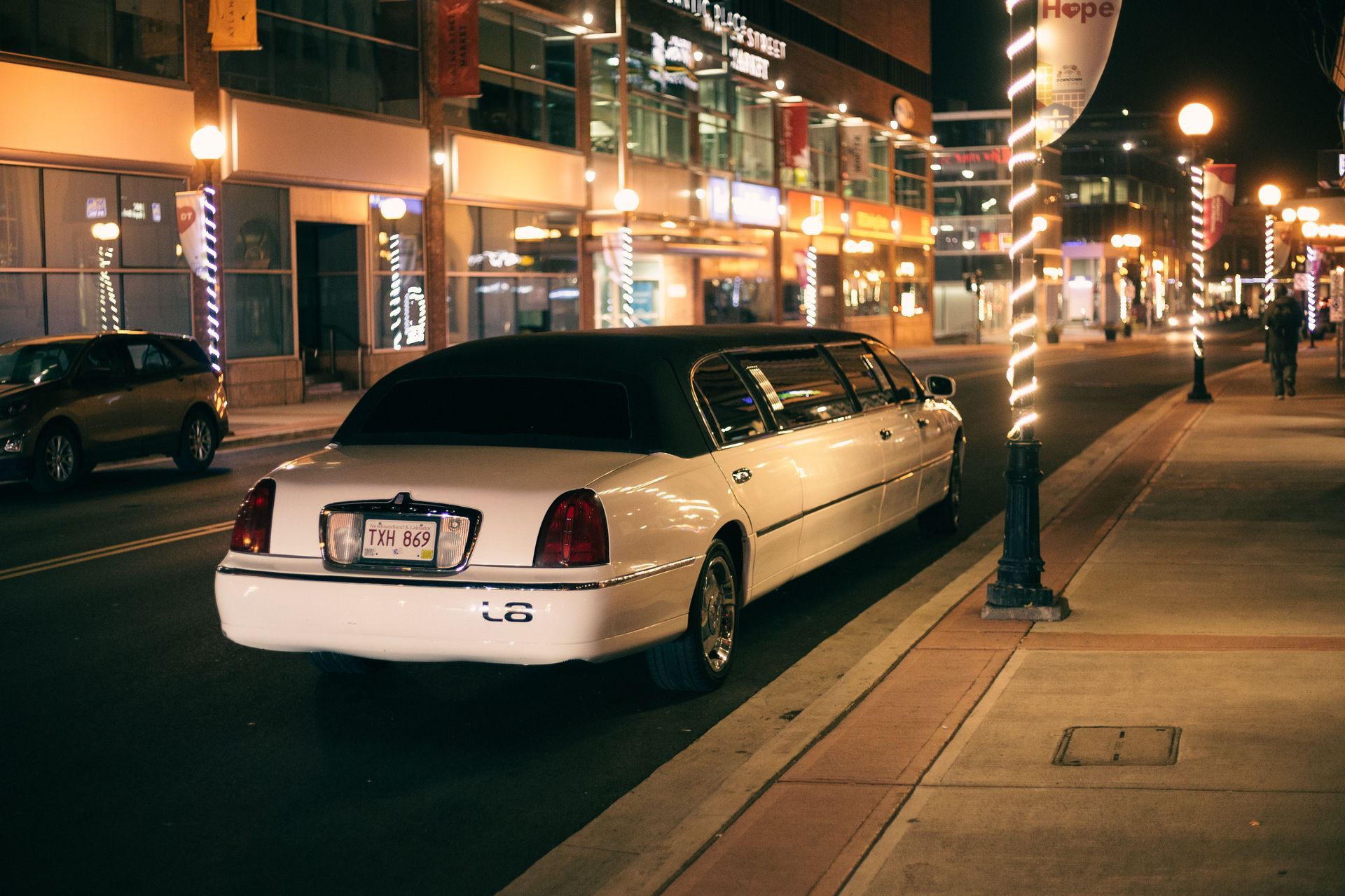 White limousine parked on a lit city street at night, with storefronts and pedestrians in the background