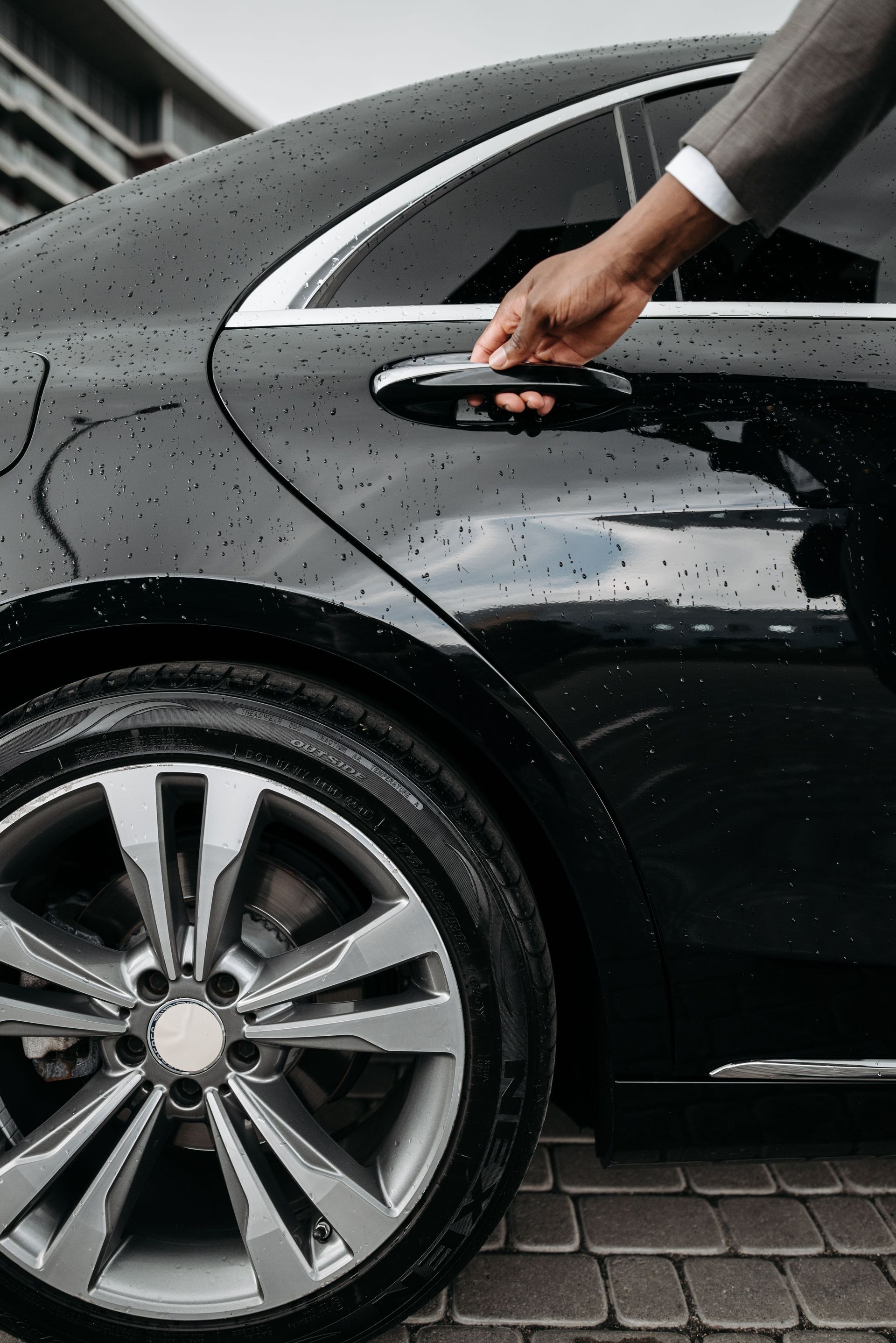 Person opening the door of a black car in the rain, with water droplets on the vehicle.