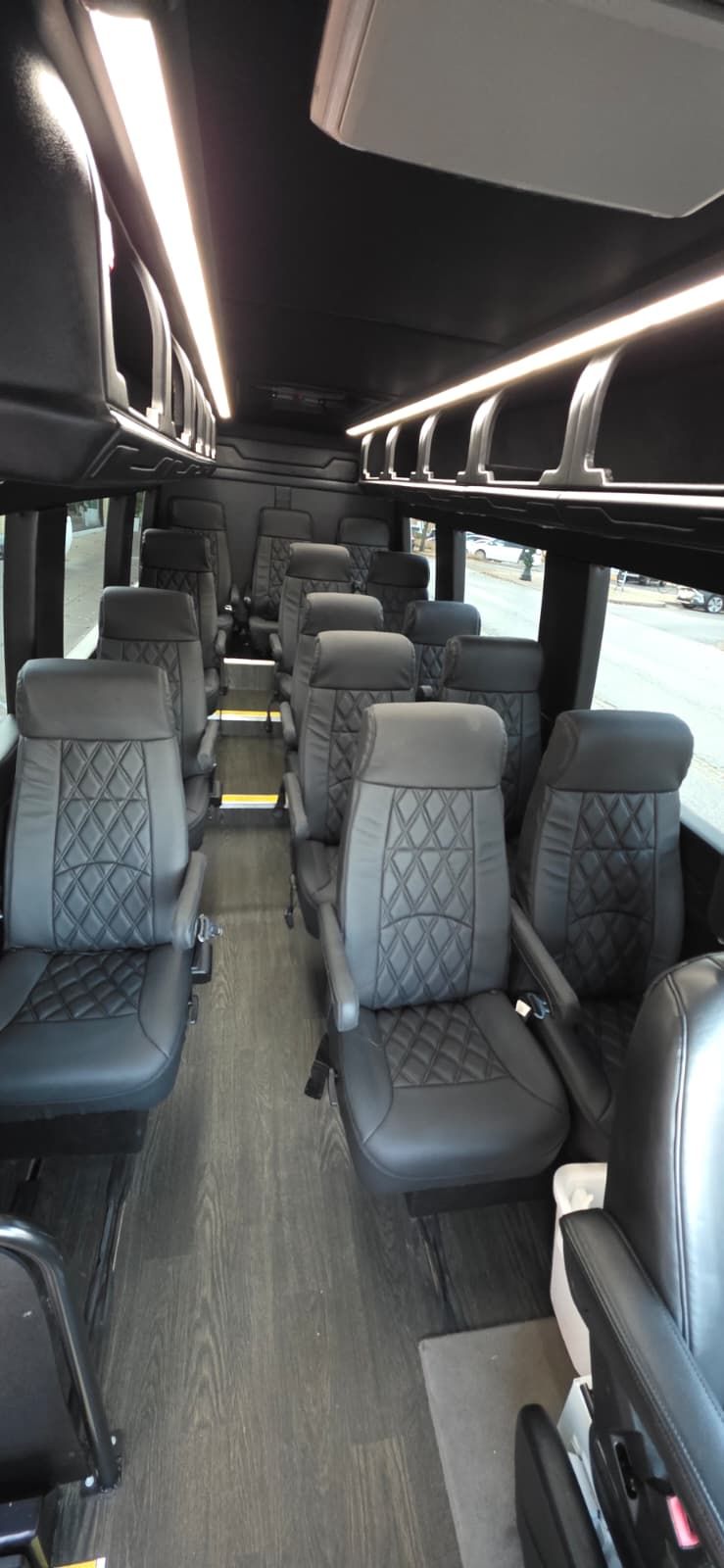 Interior of a bus, rows of black seats, gray flooring, overhead lighting.