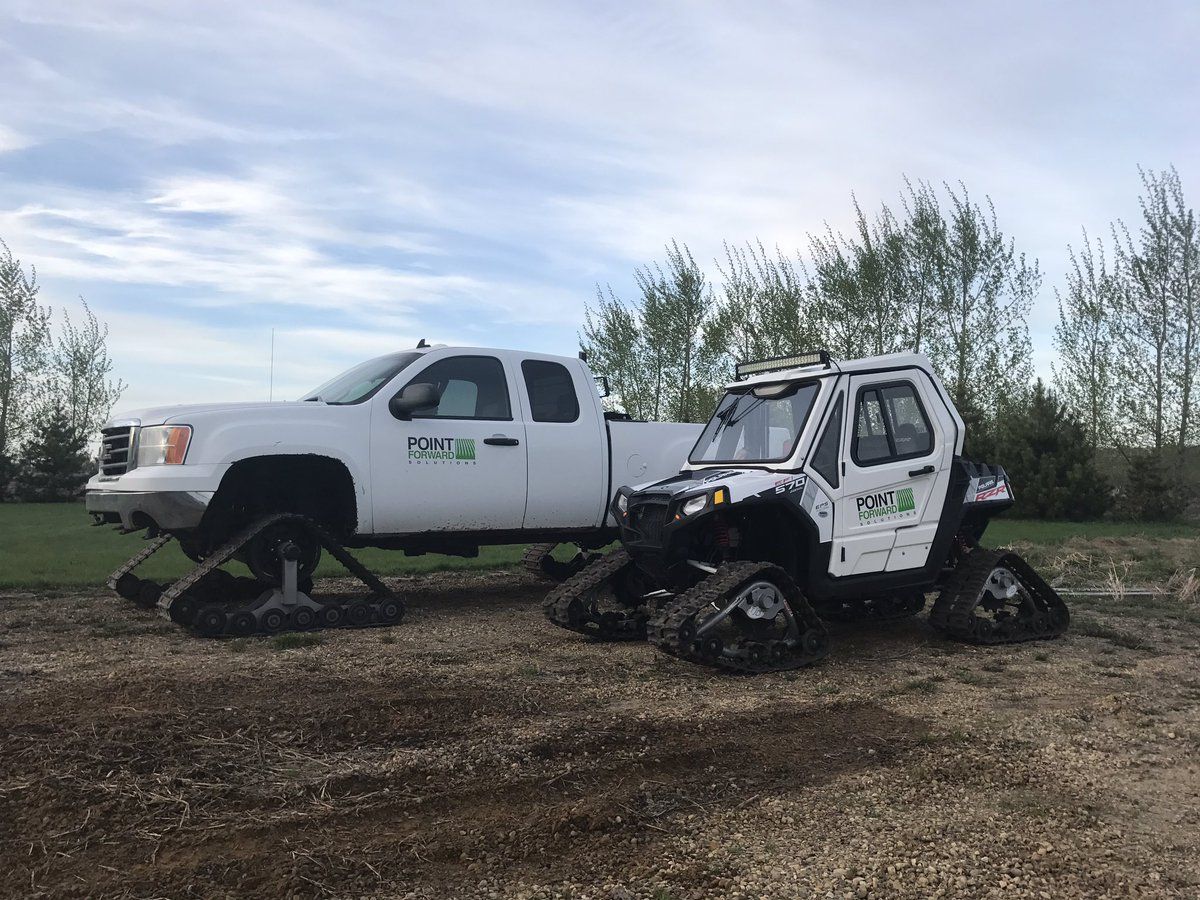 A white pickup truck and a white UTV, both equipped with tracks instead of tires, parked on a dirt lot.