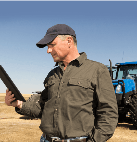 A farmer in a cap and dark shirt looks at a tablet in a field next to a blue tractor.