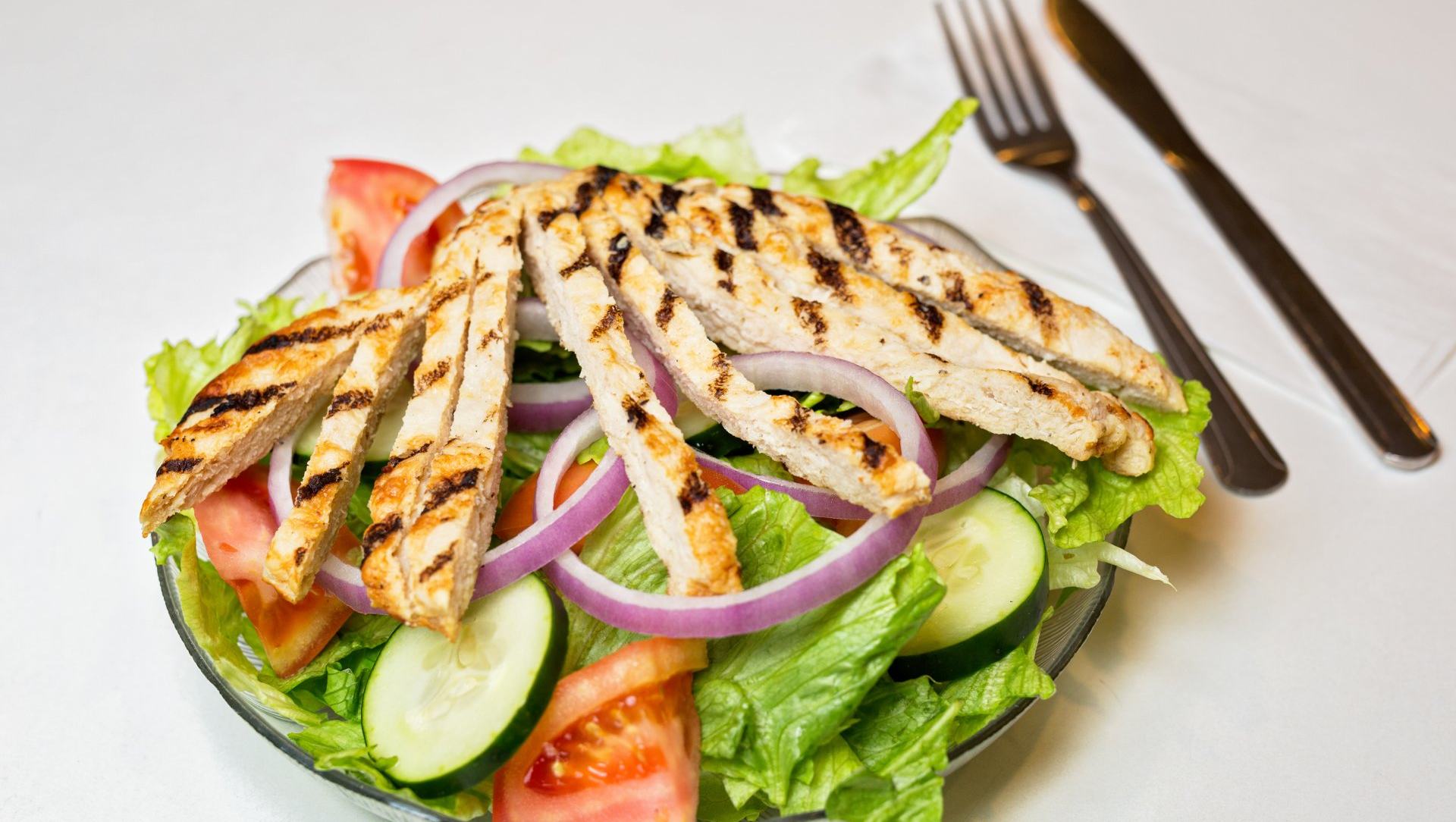 A salad with grilled chicken , lettuce , tomatoes , cucumbers and onions on a plate.