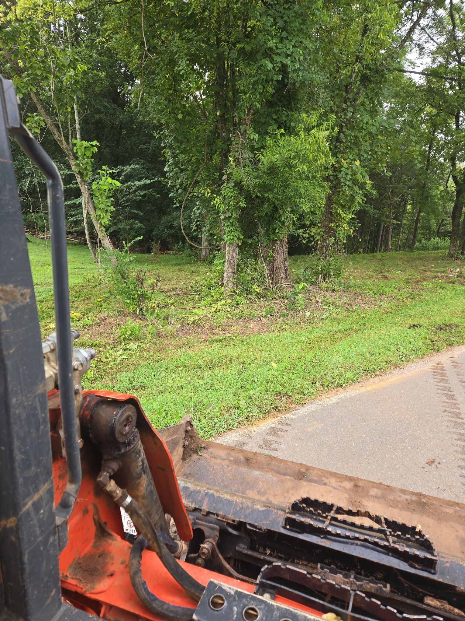 View from a skid steer of a grassy area near trees and a paved path.