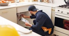 A man is kneeling down in a kitchen looking under a sink.