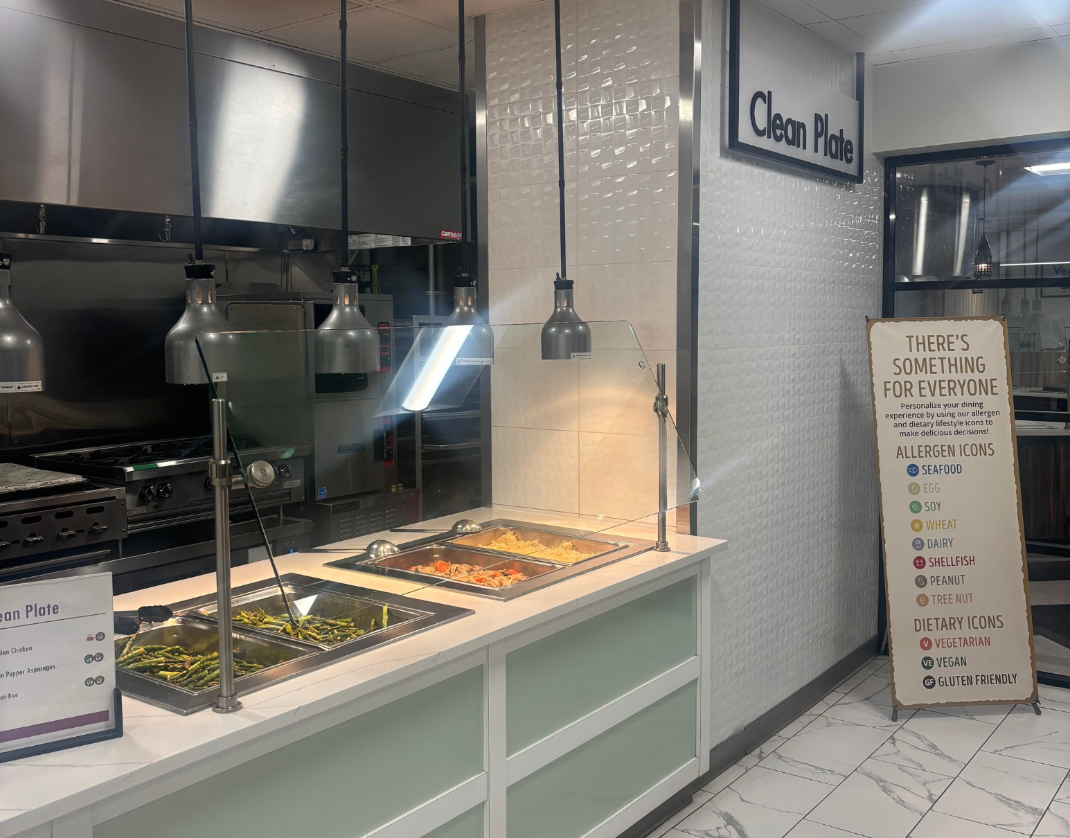 A stainless steel serving station with food trays, pendant lighting, and a menu board labeled 