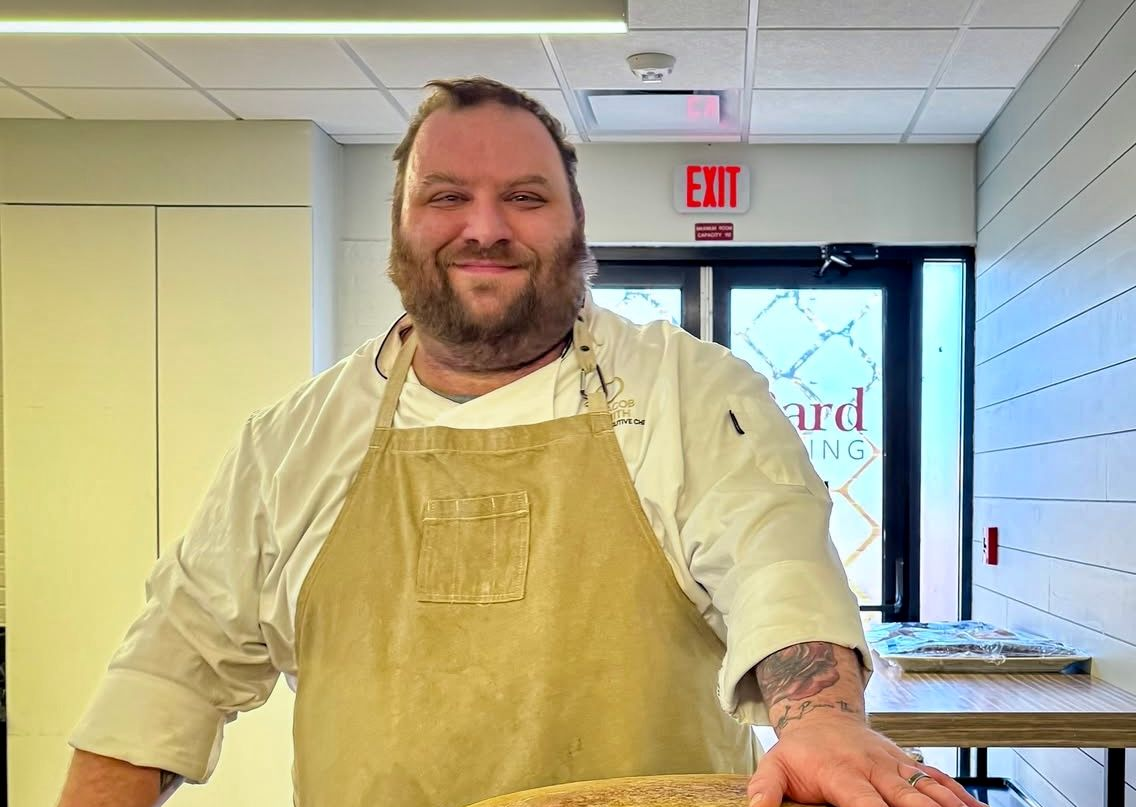 Chef in apron smiles at the camera in a kitchen setting.