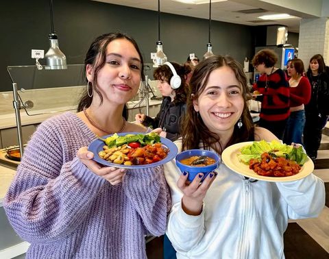 Two smiling people holding plates of food in a cafeteria. One wears a purple sweater, the other a white jacket.