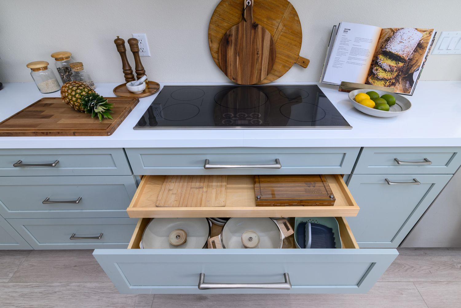 image of an induction cooktop atop  a white quartz countertop, light greenish-gray cabinets, bottom drawer open displaying pots & pans, roll out tray pulled open with cutting boards,   Rancho Conejo Neighborhood, Newbury Park, CA, open and airy kitchen remodel 
