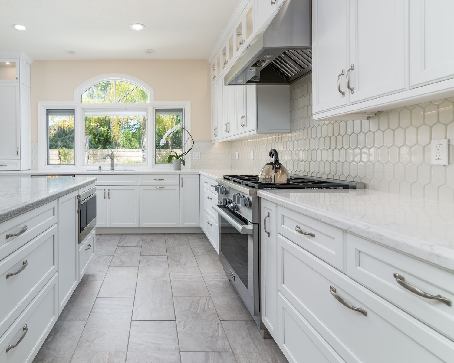 image shows the back view of a white kitchen with quartz countertops, glossy tile backsplash, stainless steel oven and hood, microwave drawer, gray hardwood floors, Newbury Park, CA, Open Concept Kitchen Expansion in Whole House Remodel