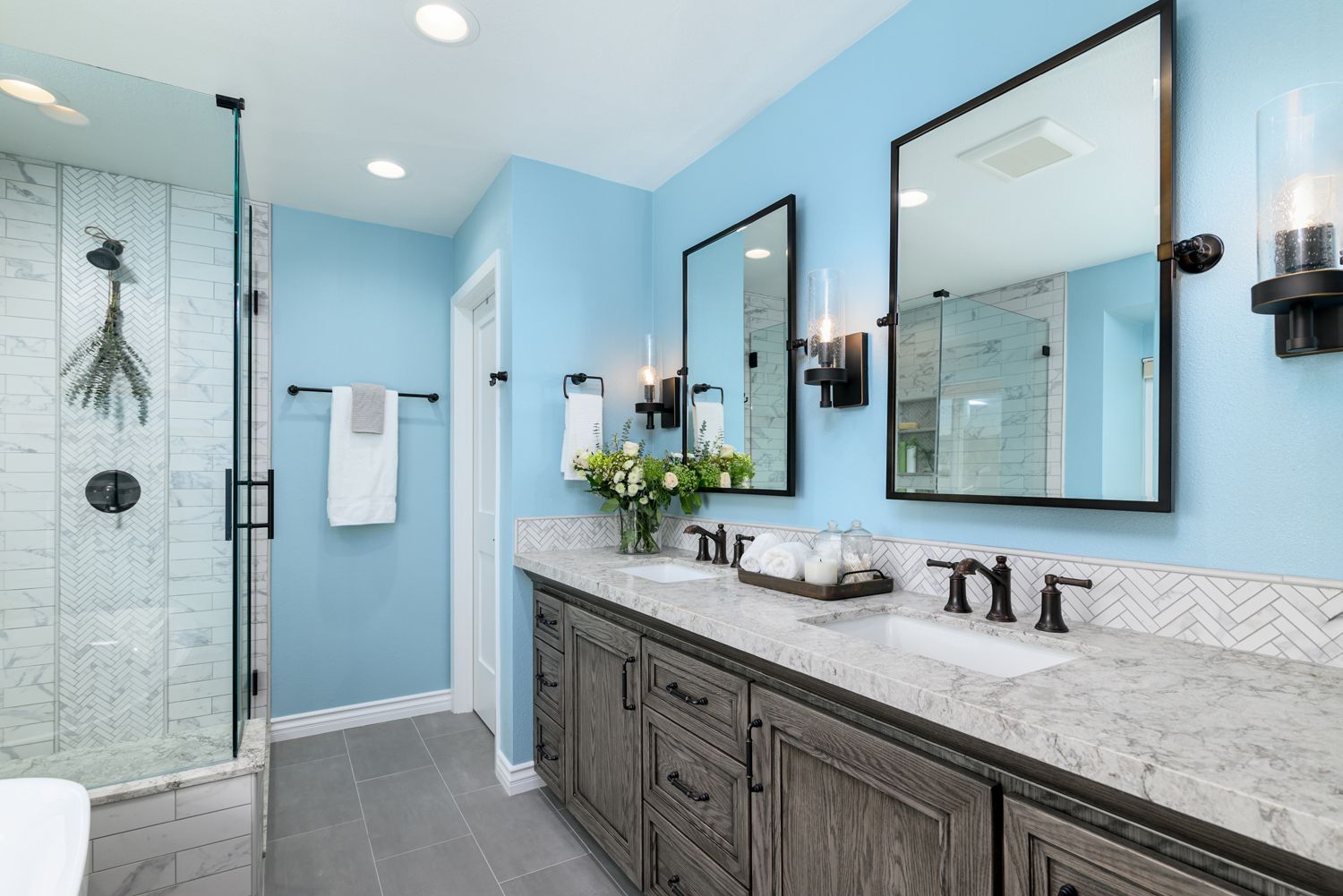 Image of a bathroom with an Omega Cabinetry vanity in oak wood w a dark gray-brown finish , a cream colored  quartz countertop by Caesarstone, sky blue walls, two sinks and two mirrors 