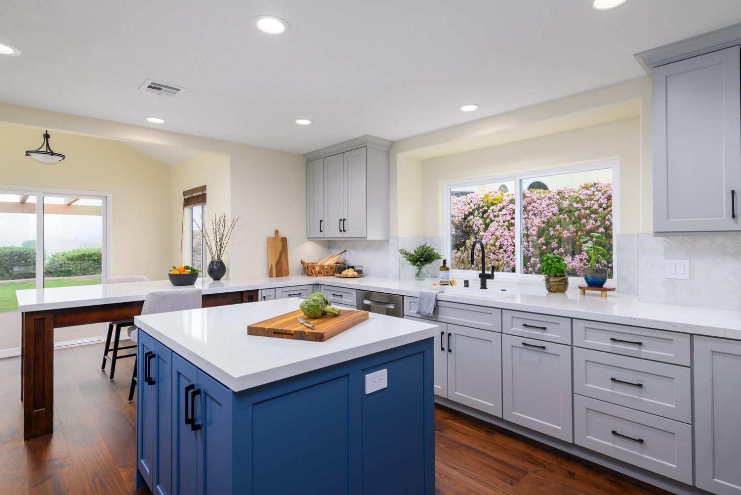 image of an attractive kitchen with a window view of pink flowers, kitchen island, rich royal blue and cool light gray cabinetry by DuraSupreme Cabinetry, white quartz countertops, dark hardwood floors, Camarillo, CA Kitchen Remodel 
