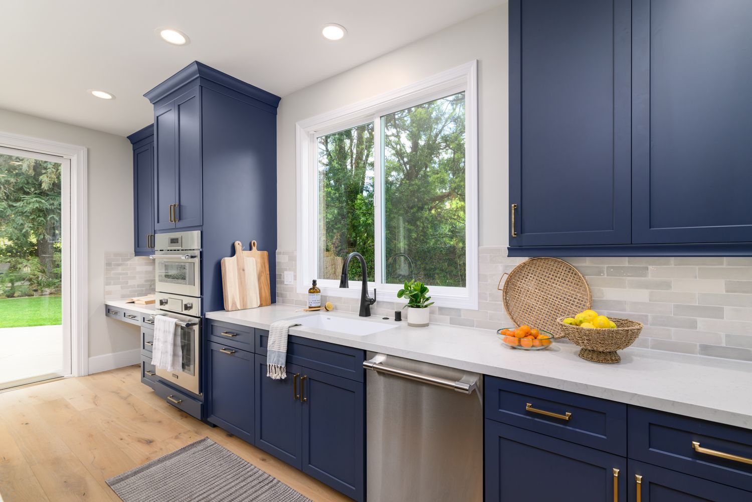 image of the back wall of a kitchen, sink, navy blue cabinetry by Omega Full Access, white quartz countertops, light gray and white subway tile backsplash, stainless steel Thermador dishwasher and double oven, new hardwood floors French Oak, Milgard Window, Agoura Hills, CA, modern style Kitchen Remodel