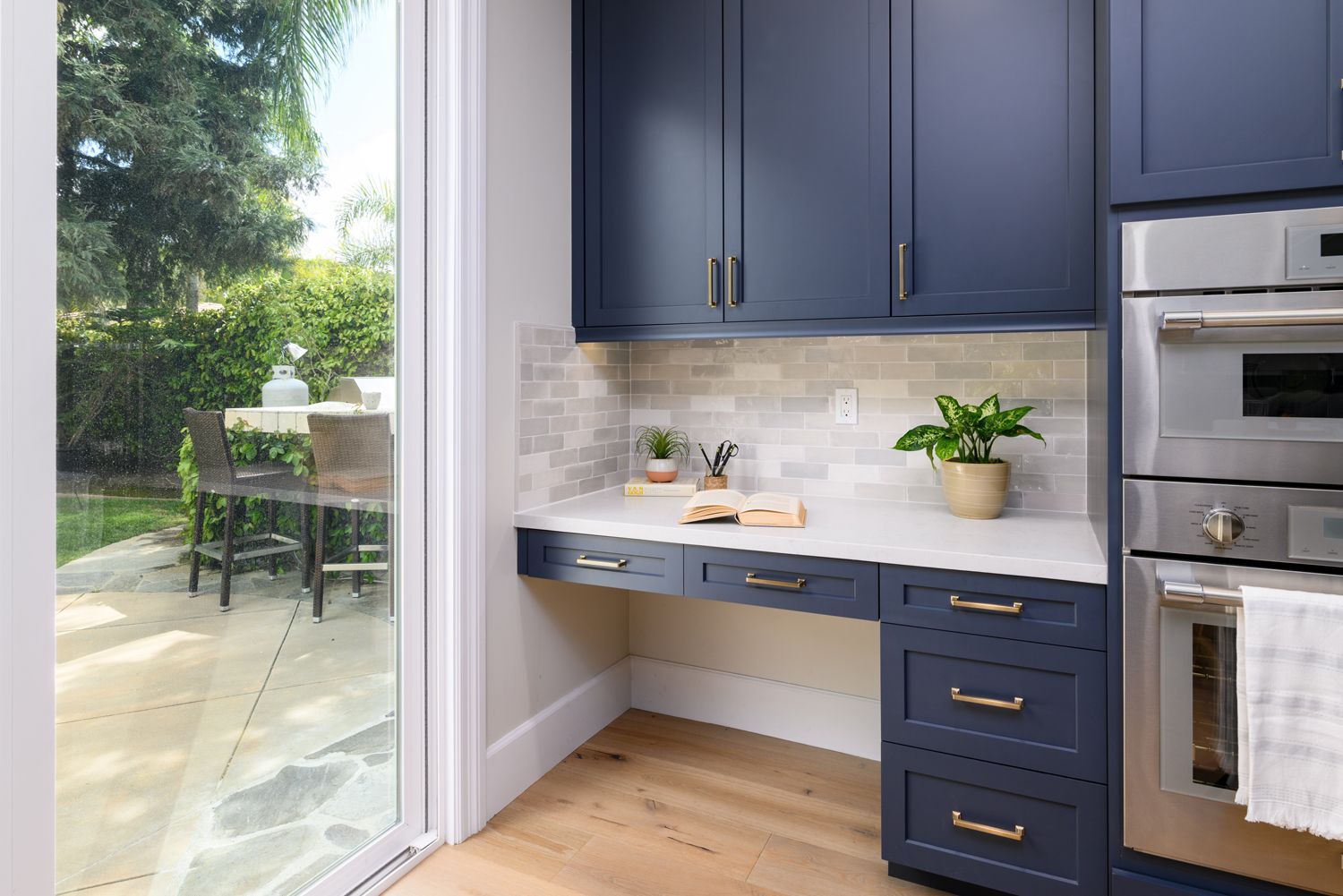 image of a built-in desk in a kitchen with three dark blue drawers, dark blue upper cabinets, Omega Full Access Cabinetry, white quartz countertops, light gray and white subway tile backsplash, new hardwood floors French Oak, Milgard sliding back door, Agoura Hills, CA, modern style Kitchen Remodel