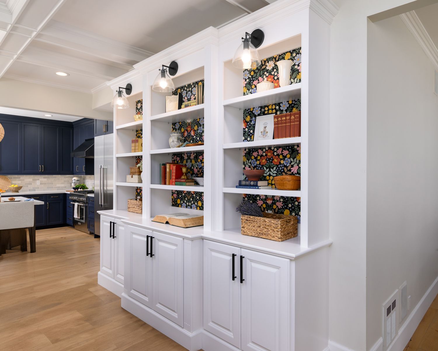 Image of a bookcase with a kitchen in the background, family room bookcase made with white painted cabinets, floral print wall paper in back of the shelves, three cupboards on the bottom, Agoura Hills, CA kitchen remodel and bookcase cabinetry remodel