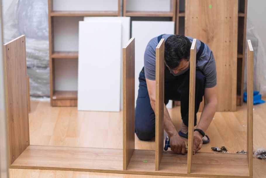 Person assembling a wooden cabinet indoors with tools and shelving parts around them