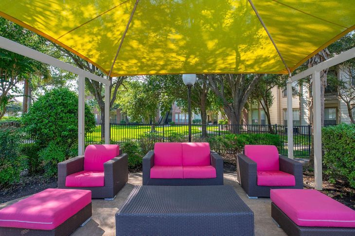 Outdoor seating area: pink cushions, yellow shade, gray furniture, surrounded by greenery.