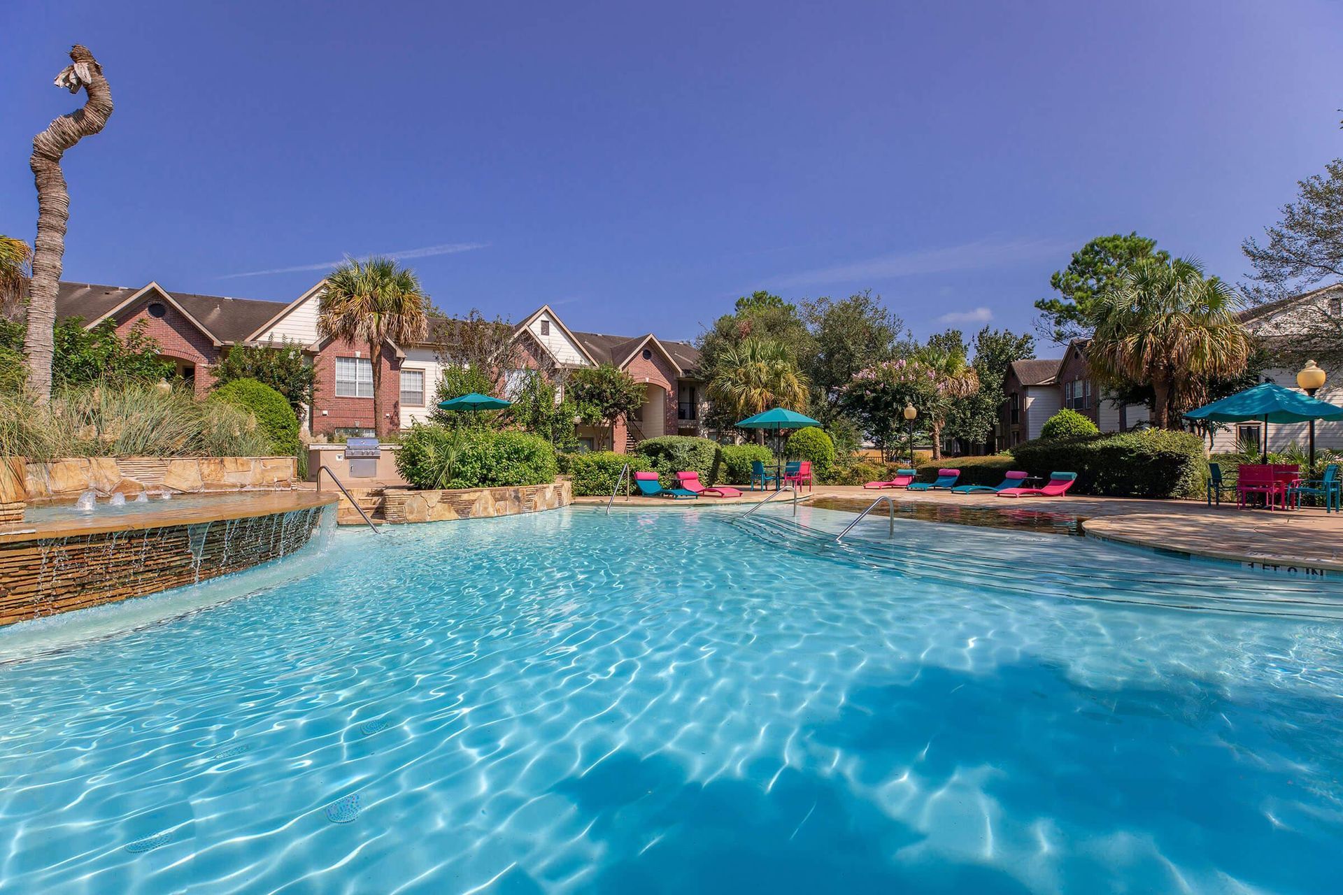 Pool with lounge chairs, umbrellas, and buildings in the background under a blue sky.