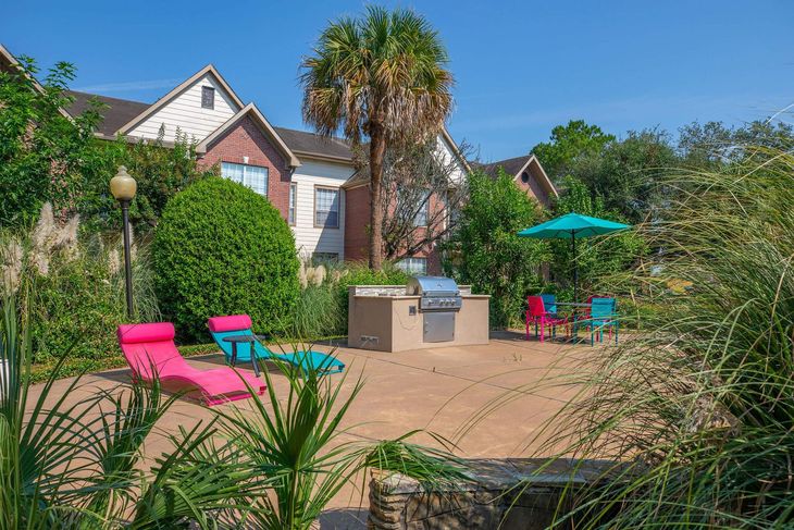 Outdoor patio with grill, lounge chairs, table, and umbrella near a building with trees.