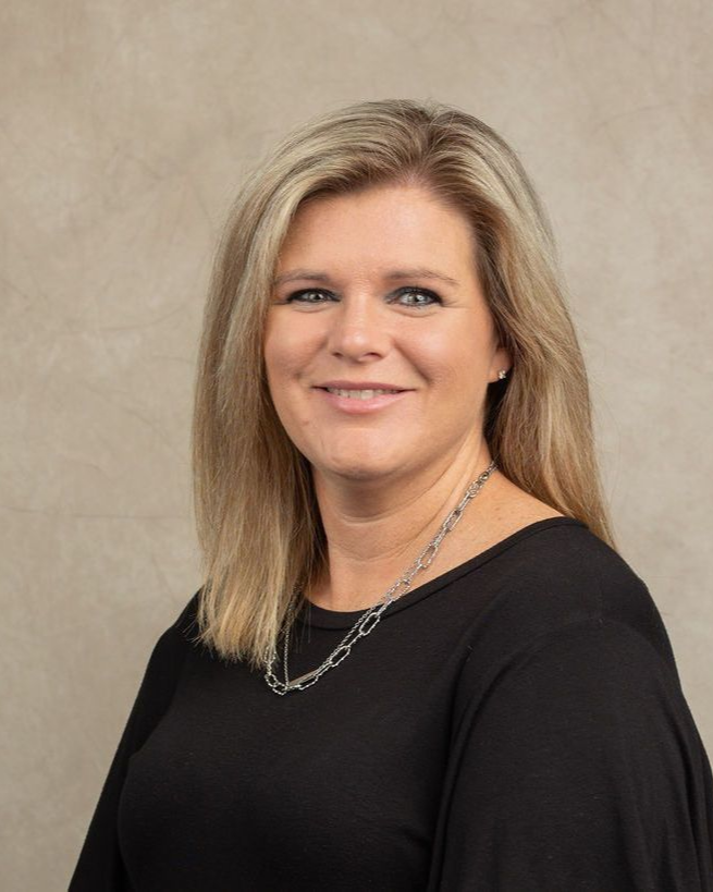 Woman with blonde hair wearing a black top and silver necklace, smiling, against a neutral background.