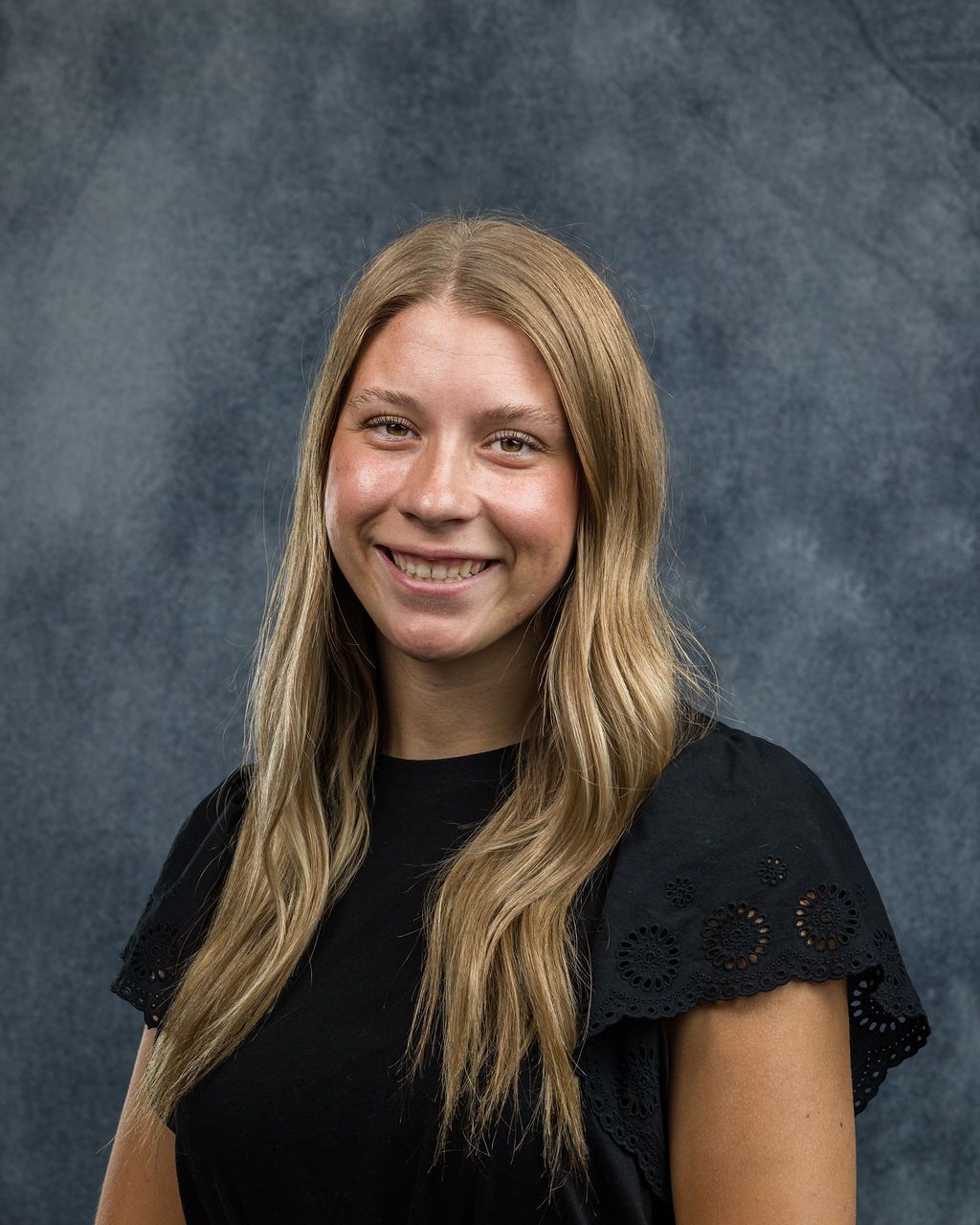 Woman with long blonde hair smiles, wearing a black top against a dark blue background.