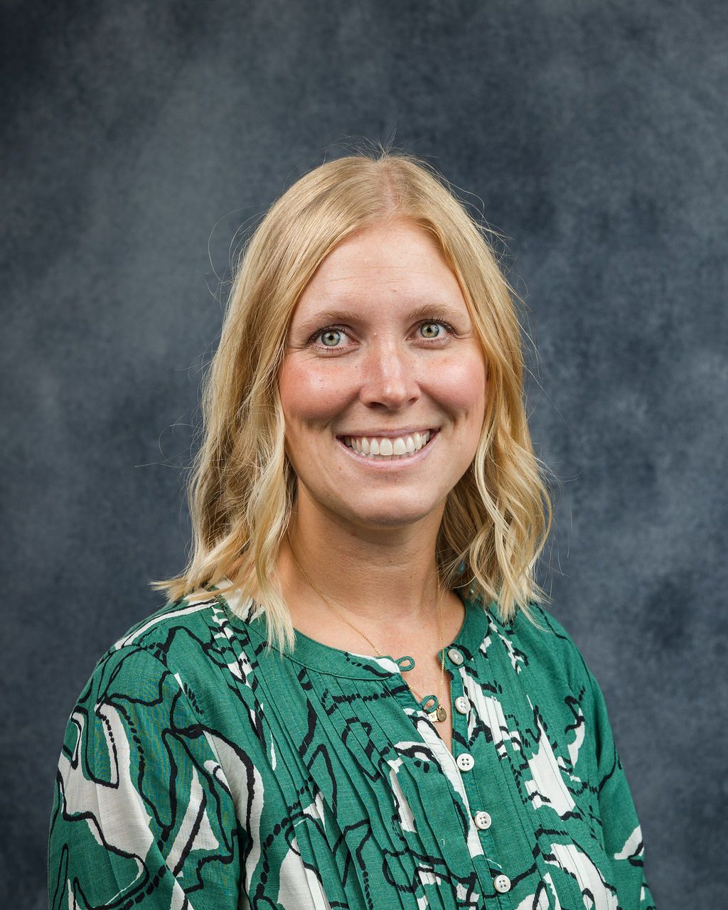 Woman with blonde hair smiling, wearing a green patterned shirt, against a dark blue background.