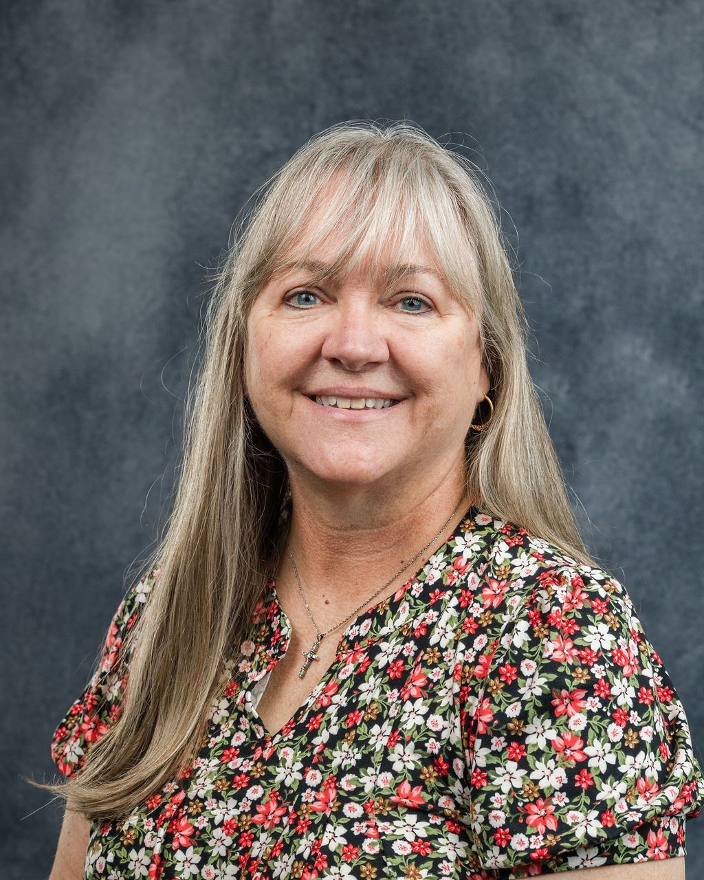 Woman with blonde hair, wearing a floral top, smiling at the camera against a grey background.