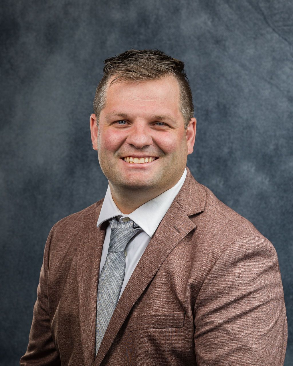 Man in a brown blazer, white shirt, and patterned tie smiles against a gray background.
