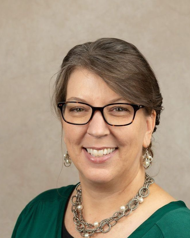Woman with glasses and a green shirt smiles at the camera, wearing jewelry, against a neutral background.