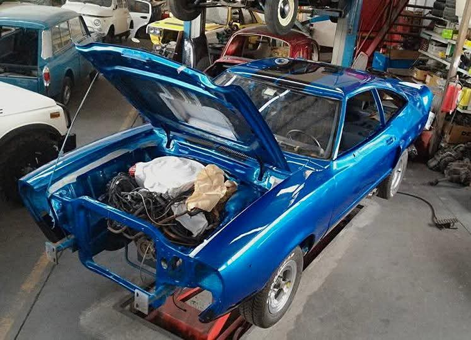 Auto d'epoca blu in riparazione in un garage, con il cofano aperto e su un ponte sollevatore.