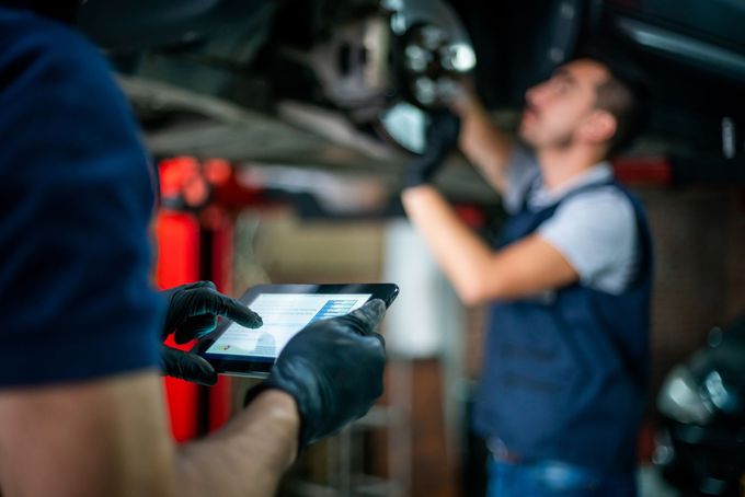 Meccanici in officina, uno usa un tablet, l'altro lavora sul telaio dell'auto.