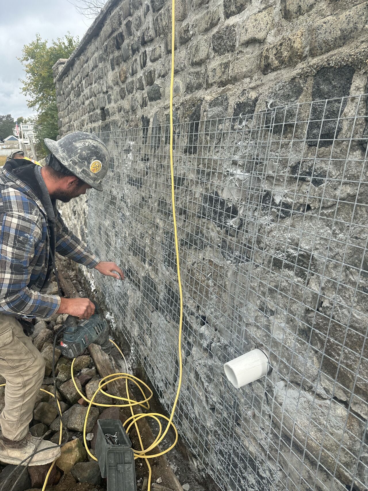 A worker in camo hard hat secures wire mesh to a stone wall with a tool; electrical cords visible.